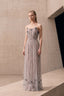 A model stands on a concrete platform wearing a strapless floor-length gown in a minimalist architectural setting. The dress is a muted lavender-gray tulle with dark floral embroidery and a thin metallic waistband.