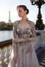 A woman wearing a long-sleeved gray-beige colored evening gown with intricate silver-pink beaded floral embroidery, standing on a Parisian bridge with the Eiffel Tower and river in the background.